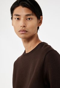 Brauner gerippter Sweatshirt mit Rundhals. Das Model schaut leicht nach links. Glatte Textur, taillierter Schnitt und subtile Nähdetails sind sichtbar.