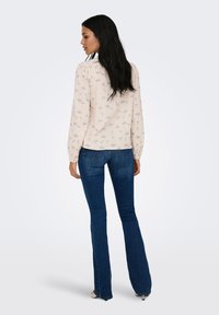 Femme aux longs cheveux noirs portant un chemisier crème à motif floral et un jean évasé bleu foncé, debout de profil sur un fond uni.