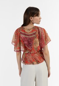 Gennemsigtig rød bluse med flutterærmer, der har guld paisley- og leopardmønstre; letvægtsstof, v-hals og afslappet pasform.