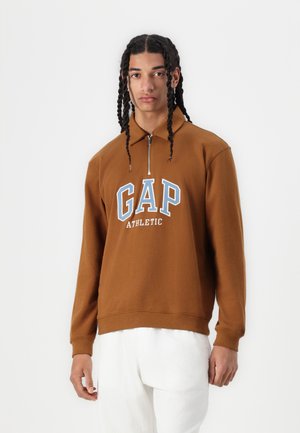 Sweat à fermeture éclair marron avec des lettres blanches et bleues "GAP ATHLETIC". Fabriqué en matériau doux avec des poignets et un ourlet côtelés.