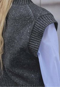 Grauer, gesprenkelter, gestrickter ärmelloser Pullover mit hohem, geripptem Kragen. Getragen über einem hellblauen, gestreiften Hemd mit glatter Textur.