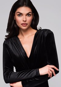 Robe en velours noir avec un décolleté en V profond et des manches longues, présentant des détails plissés aux épaules. Les mains du mannequin sont jointes devant.