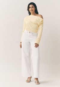 Haut jaune à manches longues et épaules dénudées, associé à un jean blanc à jambes larges. Sandales avec détails métalliques. Tissu lisse à coupe décontractée.