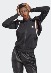 Haut de sweat à zip noir avec un design à épaules dénudées, mettant en avant des rayures blanches, le logo Adidas et des poignets côtelés. Assorti à un pantalon noir brillant.