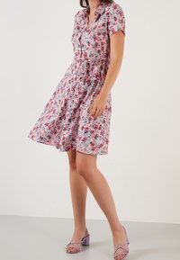 Růžové květinové šaty s krátkým rukávem, zapínání na knoflíky a stažený pas. Doplněné světle fialovými sandály na tenkých podpatcích na neutrálním pozadí.