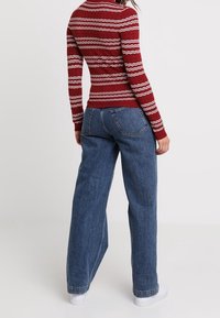 Rode long sleeve top met een wit golfpatroon, gecombineerd met wijde blauwe denim jeans. Model draagt witte sneakers. Witte achtergrond.