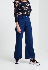 Denimowe spodnie z wysokim stanem w ciemnoniebieskim kolorze, o szerokich nogawkach i dwóch kieszeniach z przodu, połączone z czarną, kwiatową bluzką z długim rękawem oraz trampkami.