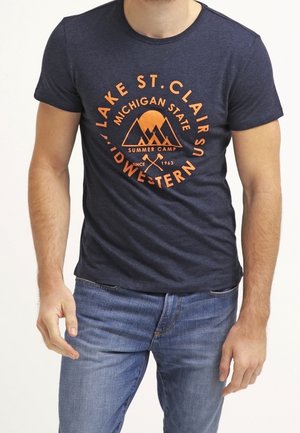Homme portant un t-shirt bleu marine avec le texte orange « Lake St. Clair Michigan State Summer Camp » et un motif de montagne, associé à un jean bleu.