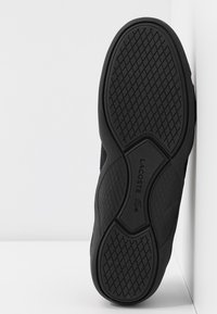 Baskets noires avec une semelle en caoutchouc texturée, présentant un motif en chevrons et un logo embossé. La tige est en tissu respirant.