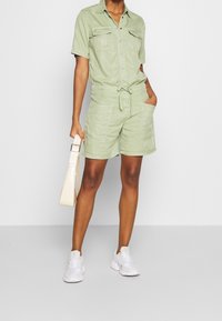 Personne portant une combinaison à manches courtes vert clair avec des poches, tenant un sac à main beige et portant des baskets blanches.