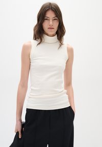 Top senza maniche a collo alto a coste in off-white, caratterizzato da un design aderente e una texture liscia, abbinato a pantaloni neri a vita alta.
