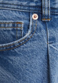 Jeans de mezclilla azul desgastados con un remache de cobre, costuras amarillas y un bolsillo con bordes deshilachados, que muestran un diseño clásico.