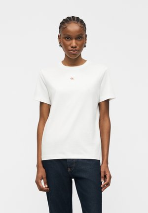 Γυναίκα με πλεκτά μαλλιά που φοράει εφαρμοστή λευκή κοντομάνικη μπλούζα με μικρό κόκκινο λογότυπο "CK" και σκούρα μπλε τζιν, σε ουδέτερη στάση.