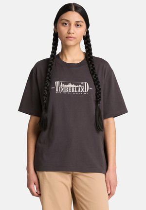 Femme aux longs cheveux tressés portant un t-shirt Timberland foncé et un pantalon beige, debout devant un fond clair uni.