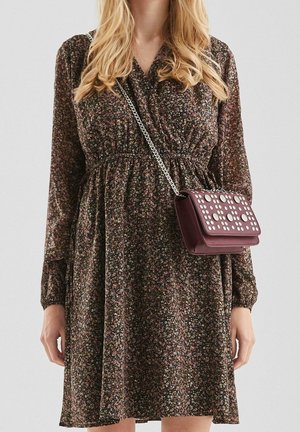 Kvinde iført en langærmet mørk blomsterkjole med en indsnævret talje, bærende en lille mørkerød crossbody-taske med sølvnitter og kædremme.