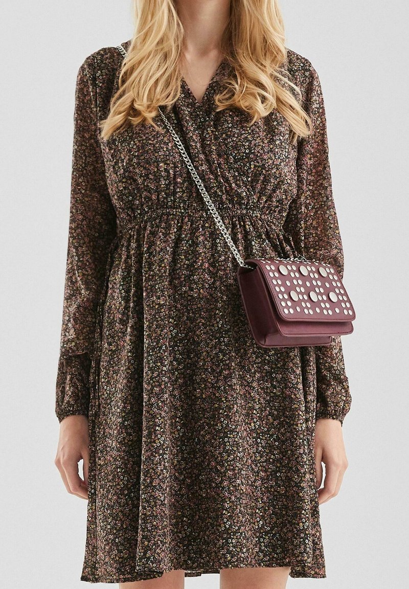 Femme portant une robe longue à manches longues à motif floral foncé avec une taille froncée, portant un petit sac bandoulière bordeaux avec des clous argentés et une bandoulière en chaîne.