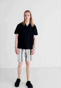 Juodas trumpomis rankovėmis marškinėliai su mygtukais, dryžuotos baltos ir mėlynos šortai, ir juodos slip-on bateliai. Paprasta fono, neutraliomis tonomis.