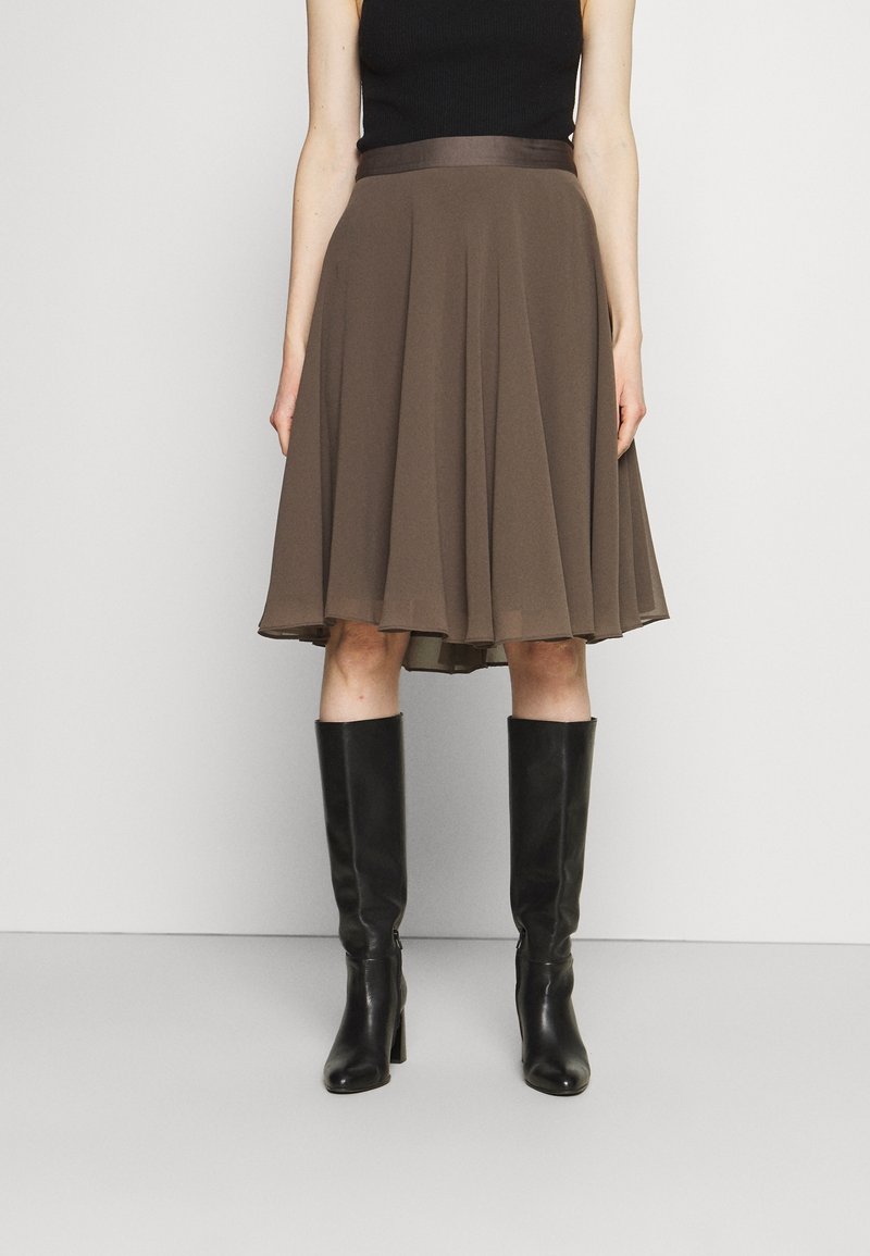 Femme portant une jupe marron évasée jusqu'aux genoux, un haut noir sans manches et des bottes en cuir noires montantes, debout devant un fond uni.