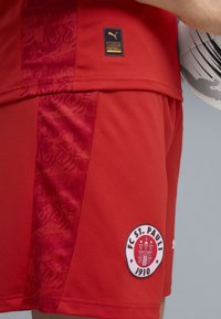 Röda fotbollsshorts i andningsbart material med ett strukturerat mönster, Puma-logotyp och FC St. Pauli-märke.
