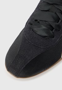 Μαύρο αθλητικό παπούτσι από μπροστά, κοντινή λήψη που δείχνει το λείο ύφασμα στο μπροστινό μέρος, το πάνελ από σουέντ και τα πλατιά κορδόνια από σατέν σε ανοιχτό φόντο.