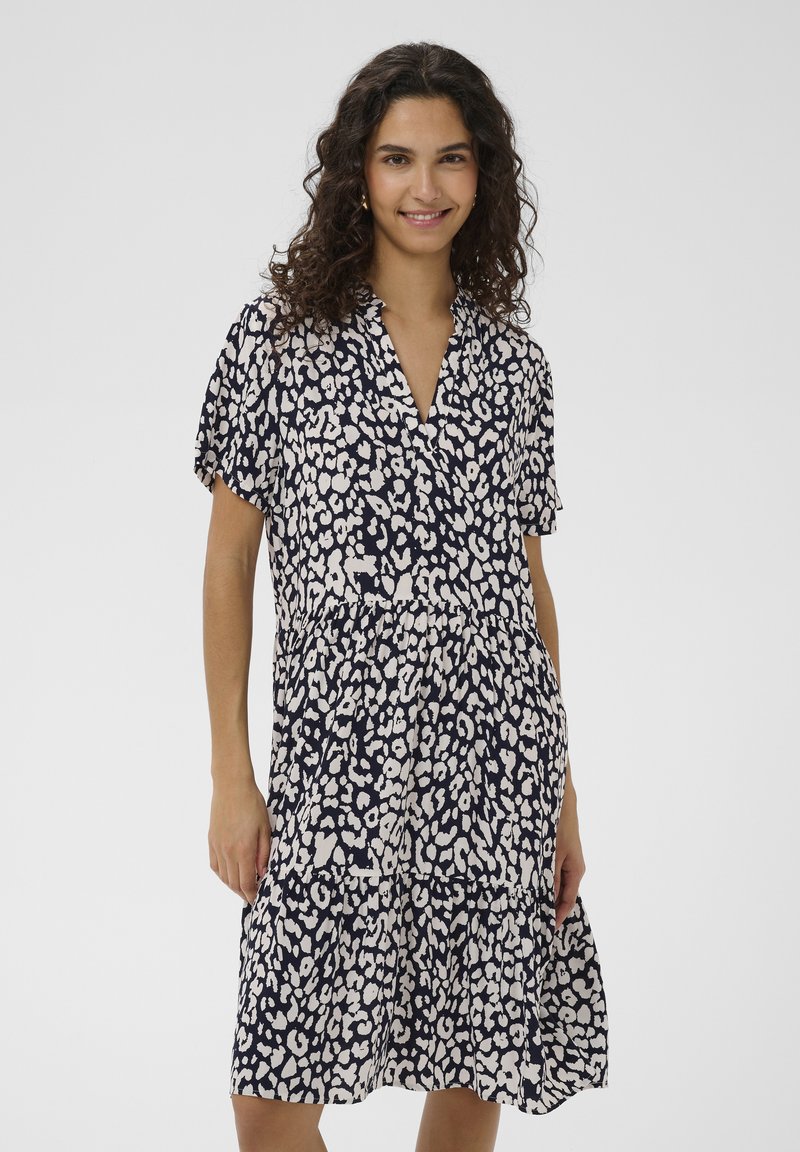 Femme souriante portant une robe léopard noire et blanche mi-longue avec des manches courtes et un col en V.