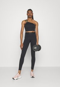 Top corto negro de un solo hombro con un acabado texturizado y una tira estampada, combinado con leggings negros de cintura alta y zapatillas deportivas rosa claro.