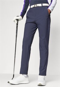 Navy Golfhosen mit glatter Textur, ausgestattet mit Fronttaschen und Gürtelschlaufen, kombiniert mit weißen Schuhen und einem Golfschläger in der Hand.