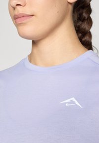 Hele lilla sportiven shirt ümmarguse kaelusega. Vasakul rinnal on valge Nike'i logo aktsent. Siledat, keha ümber oleva kangaga tekstuuri.