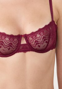 Bordowy koronkowy stanik ma przezroczyste miseczki z geometrycznym wzorem, centralne wycięcie i falbany wzdłuż górnej krawędzi. Regulowane ramiączka.