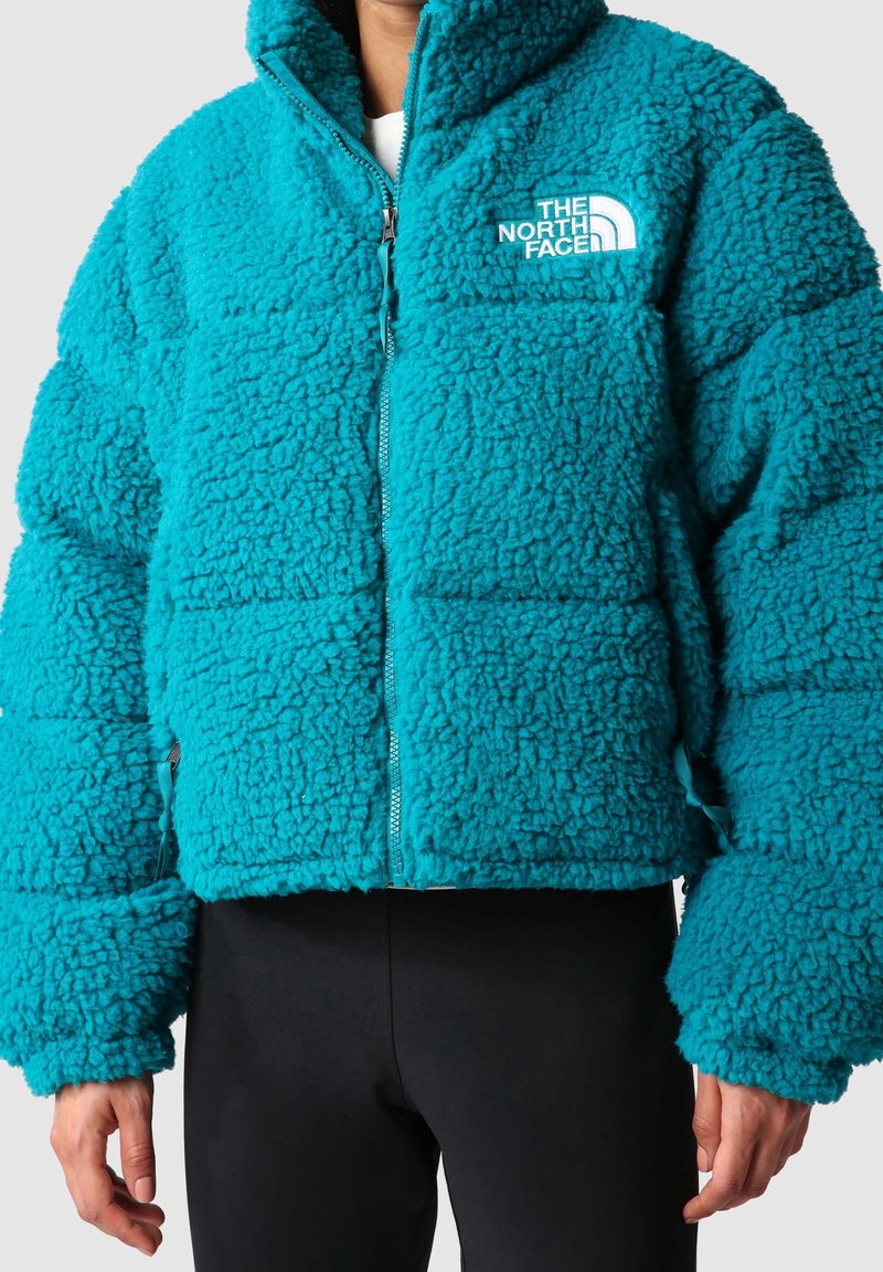 Petrolejově modrá huňatá fleecová bunda s vysokým límcem a hrubou texturou. Má celopropínací zip a logo na levé horní straně. Dlouhé, široké rukávy.