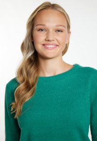 Maglione verde, morbido, con una vestibilità ampia e un collo rotondo, realizzato in un materiale testurizzato e soffice. Capelli acconciati in onde morbide.