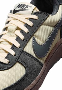 Sneaker Nike z kremowym materiałowym wierzchem, szarymi zamszowymi i skórzanymi akcentami, jasnobeżowymi sznurowadłami oraz ciemną gumową podeszwą z detalem logo.