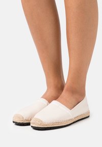 Espadrilles en toile beige avec une semelle en jute tressé, arborant un logo blanc sur le dessus, un design plat et un bout arrondi.