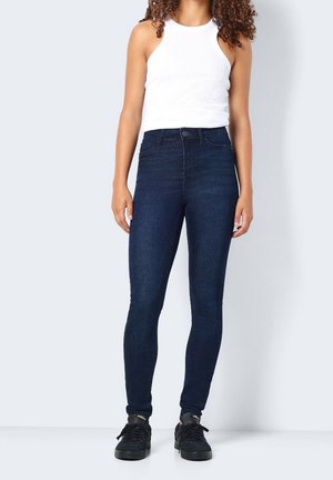 Person, die ein weißes ärmelloses Oberteil, dunkelblaue hoch taillierte Skinny Jeans und schwarze Sneakers trägt, steht vor einem schlichten Hintergrund.