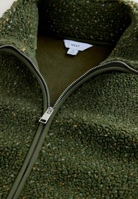 Green fleece zip-up jacket with a thick, textured surface, silver zipper, and matching collar. Label reads "NEXT" with size tag.