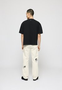 Camiseta negra de gran tamaño y pantalones cargo crema con intrincados patrones negros. Los pantalones cuentan con bolsillos laterales y un ajuste relajado. Zapatos negros.