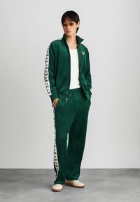 Zielony dres z weluru z zamkiem błyskawicznym i dopasowanymi spodniami, z bocznymi paskami z powtarzającym się wzorem logo. Białe akcenty i miękka tekstura.