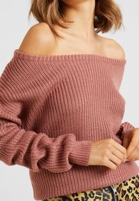 Femme portant un pull en tricot rose poussiéreux à épaules dénudées avec des manches longues et un pantalon à imprimé léopard avec une fermeture éclair à l'avant.