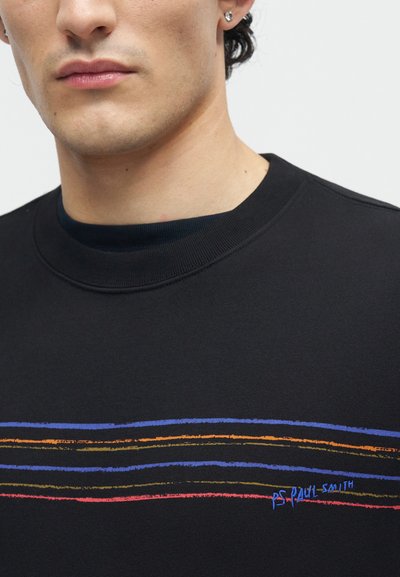 Κοντινό πλάνο ατόμου που φοράει μαύρο φούτερ με πολύχρωμες οριζόντιες γραμμές και μικρό κείμενο "PS Paul Smith" στο στήθος.