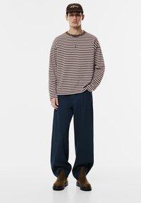 Młody mężczyzna stoi w brązowej, pasiasta koszuli z długim rękawem, granatowych szerokich spodniach, brązowych butach, brązowej czapce oraz srebrnym naszyjniku.