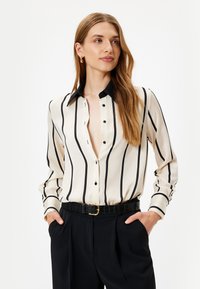 Blusa de crema con rayas verticales negras, cuello negro y frente con botones. Mangas largas con puños abotonados, combinada con pantalones negros de cintura alta.