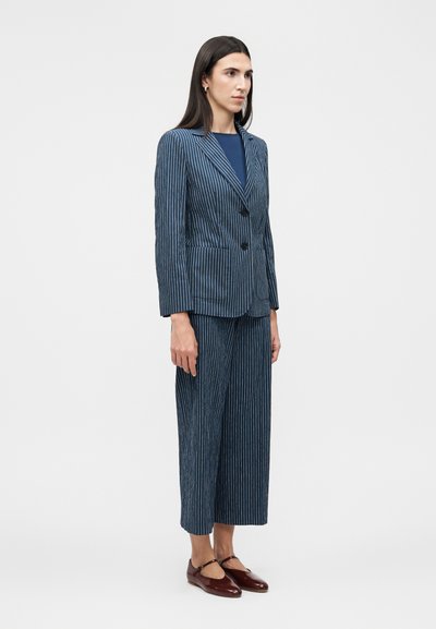 Γυναίκα όρθια, φοράει μπλε σακάκι με ρίγες και ταιριαστό παντελόνι με φαρδιά καμπάνια μέχρι τον αστράγαλο, καθώς και σκούρα κόκκινα παπούτσια Mary Jane.