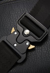 Sangle en nylon noir avec une surface texturée, dotée d'une boucle noire et dorée avec des accents en argent et un rivet en argent proéminent.