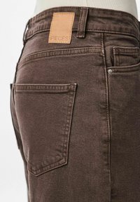 Bruna denimjeans med texturerad yta, med bakfickor och en läderlapp med texten "PIECES" vid midjan.