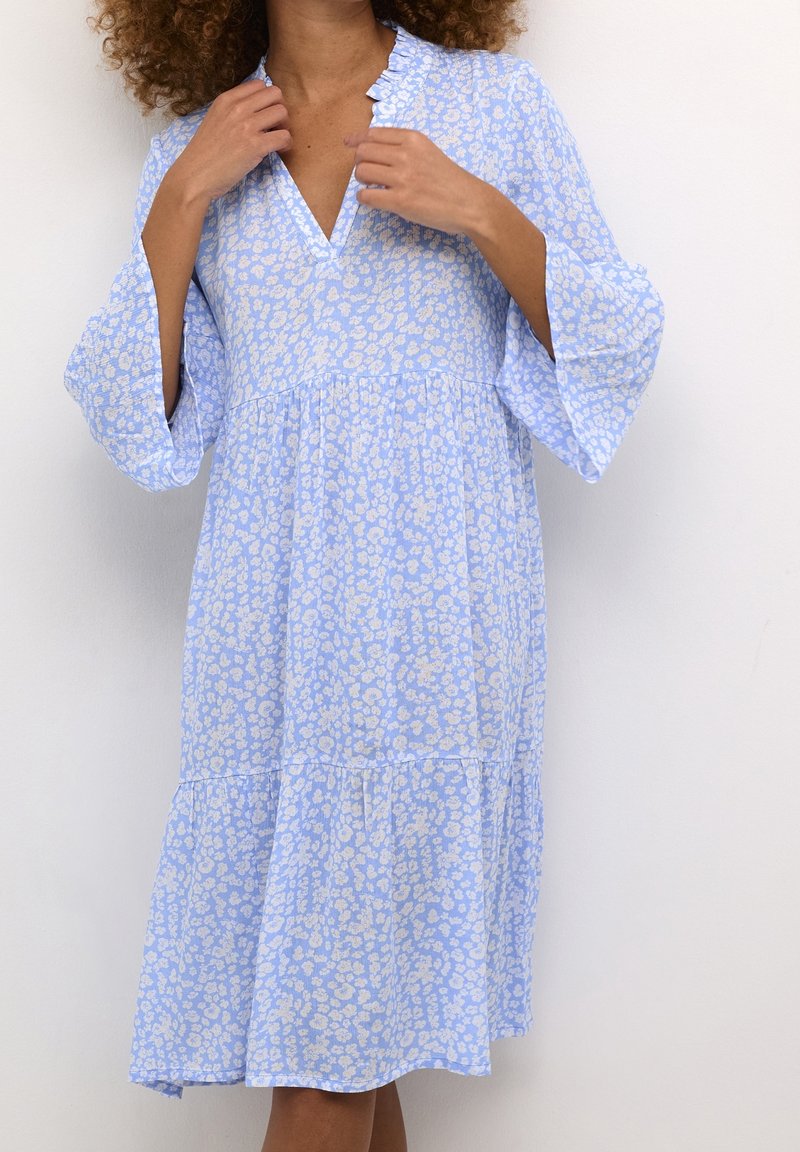 Šaty s květinovým vzorem v světlé modré a krémové barvě, s výstřihem do V, volánkovým výstřihem a zvonovými rukávy. Víceúrovňový design přidává objem.