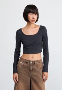 Black long-sleeve cropped top with a ribbed texture, featuring a wide scoop neckline and subtle seam detailing paired with tan high-waisted pants.
