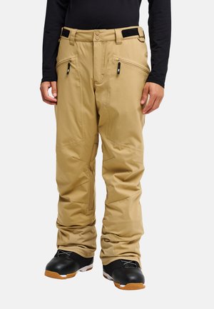 Pantalon de neige beige avec poches avant zippées, ceintures ajustables à la taille, porté avec des chaussures noires et un top noir à manches longues.