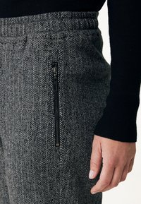 Šedé kalhoty s herringbone vzorem a kapsou na zip, vyznačující se širokým elastickým pasem a měkkou texturovanou látkou.