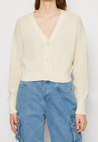 Krämfärgad virkade kofta med V-ringning, långa ärmar och knäppning fram. Har ett strukturerat stickat mönster och ribbat fåll.