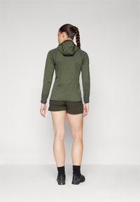 Chaqueta con capucha de tela verde oscuro con mangas largas, combinada con pantalones cortos verde oscuro. Presenta un diseño ajustado y material texturizado.